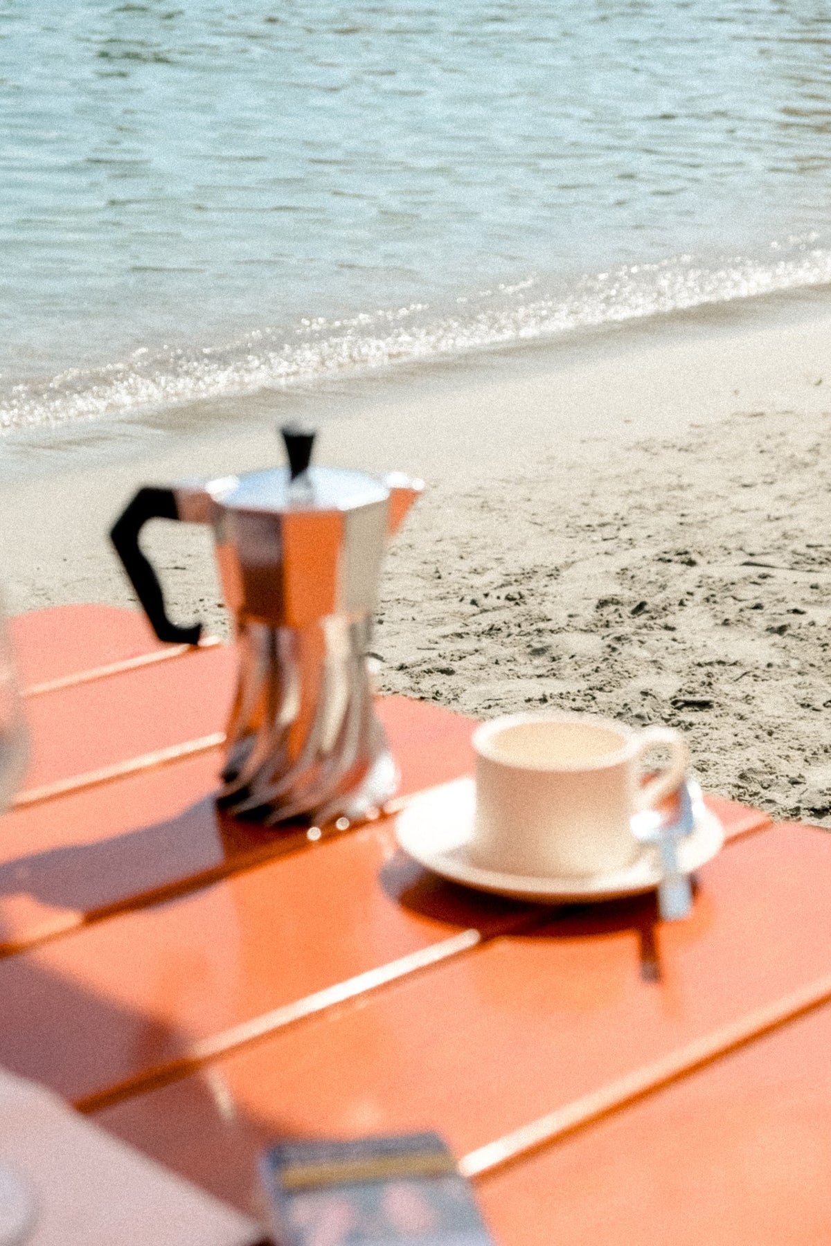 Turbo Moka 2-cup aluminium moka pot on an orange beach table beside an espresso cup, shoreline behind
