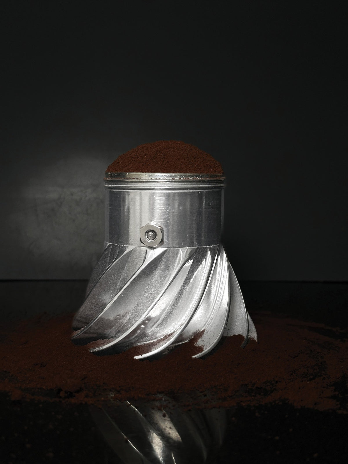 Turbo Moka boiler filled with ground coffee on a dark reflective surface scattered with coffee grounds