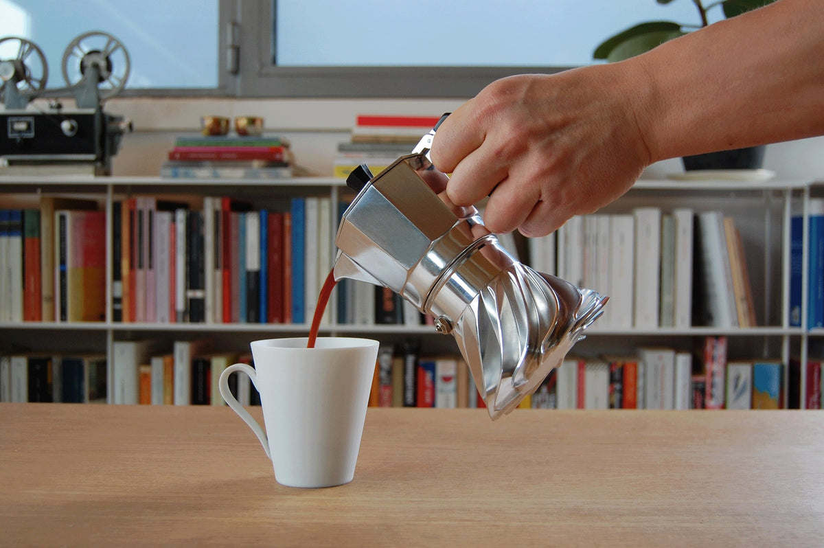 Hand pouring coffee from the Turbo Moka into a white cup in front of a bookshelf