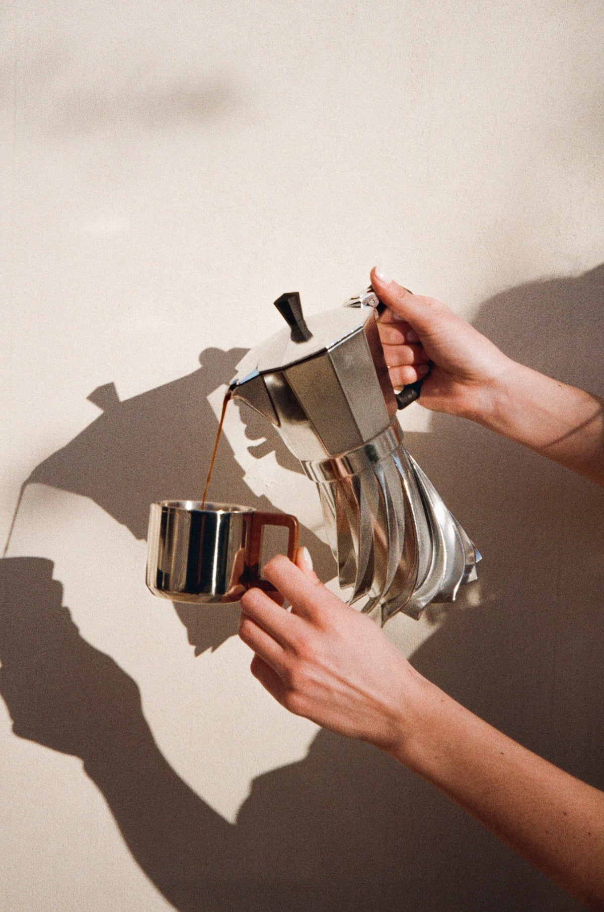 Hands pouring coffee from the Turbo Moka into a small steel cup against a warm plaster wall in hard sunlight