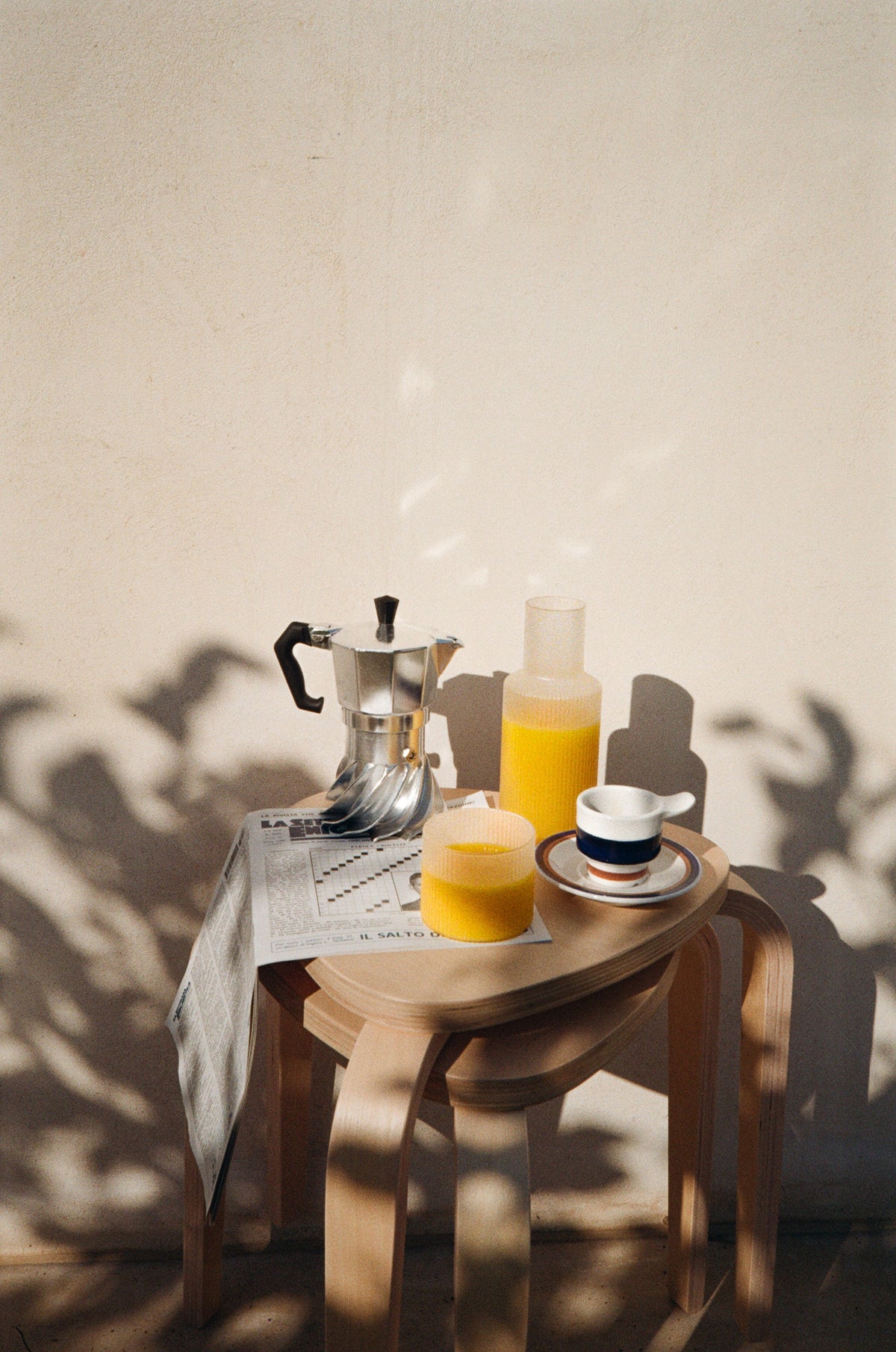 Turbo Moka moka pot on a wooden stool with a carafe of orange juice, a glass, an espresso cup and a newspaper, leaf shadows across the sunlit wall behind