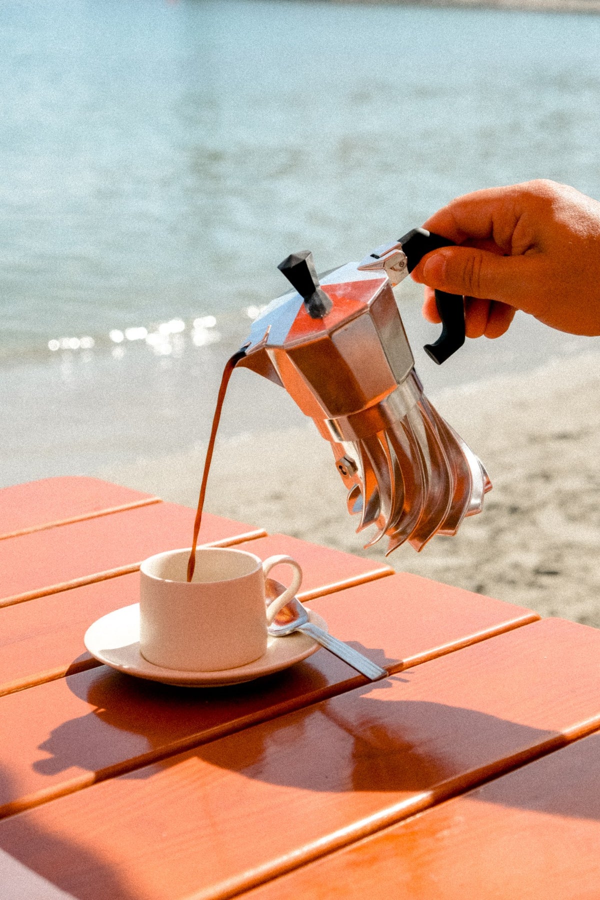 Pouring coffee from the Turbo Moka 2-cup into a white cup on an orange table by the sea