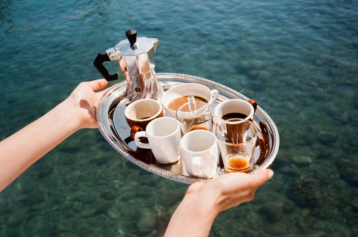 Hands holding a silver tray of espresso cups and the Turbo Moka out over clear turquoise water