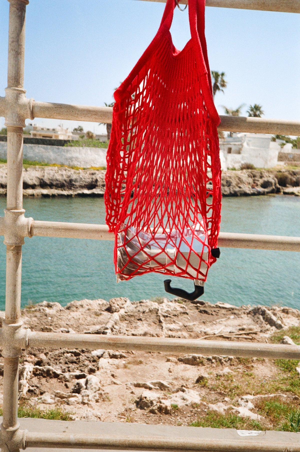 Turbo Moka carried in a red string market bag hanging from a white seafront railing, with the sea and a rocky shoreline behind